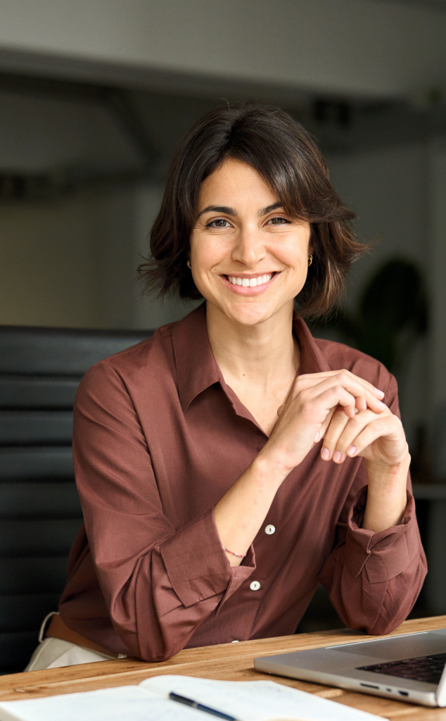 Smiling woman in a maroon blouse sits at a desk with a laptop and notebook in a modern office setting.