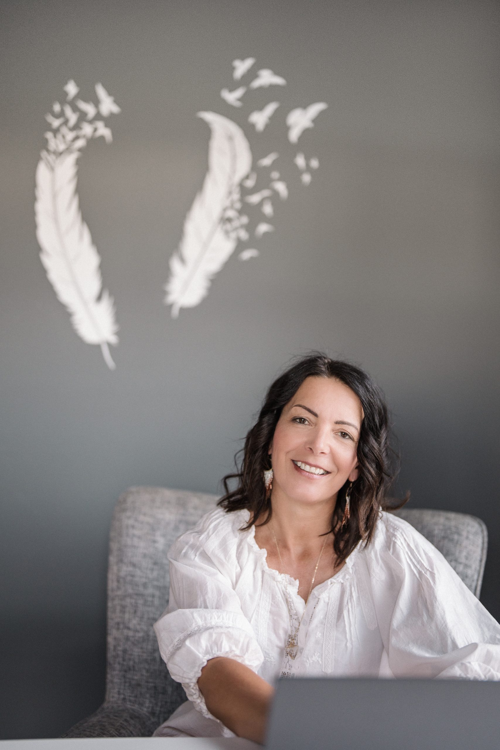 Smiling woman in a white blouse sits at a desk with a laptop, in front of a wall decorated with white feather and bird illustrations.