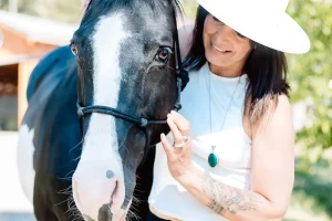 Smiling woman in a white hat and sleeveless top gently holds the bridle of a black-and-white horse outdoors on a sunny day.