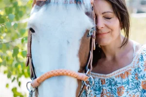 Smiling woman in a floral blouse gently rests her face against a white horse’s head, eyes closed in a moment of peaceful connection.