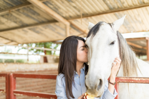 Woman in a light blue shirt kisses a white horse on the forehead while gently holding its face in a stable.